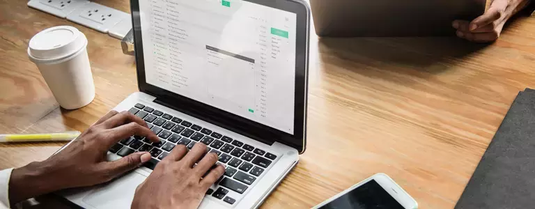 Man typing on a Macbook at work with a disposable coffee cup