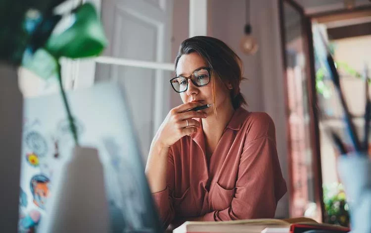 Woman focused on computer, holding pen while analyzing information.