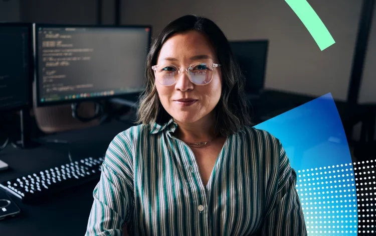 A woman with shoulder-length hair, wearing glasses and a striped blouse, poses confidently in an office setting with computer screens in the background using Proofpoint Human Risk Explorer. Soft lighting adds a warm tone to the scene.