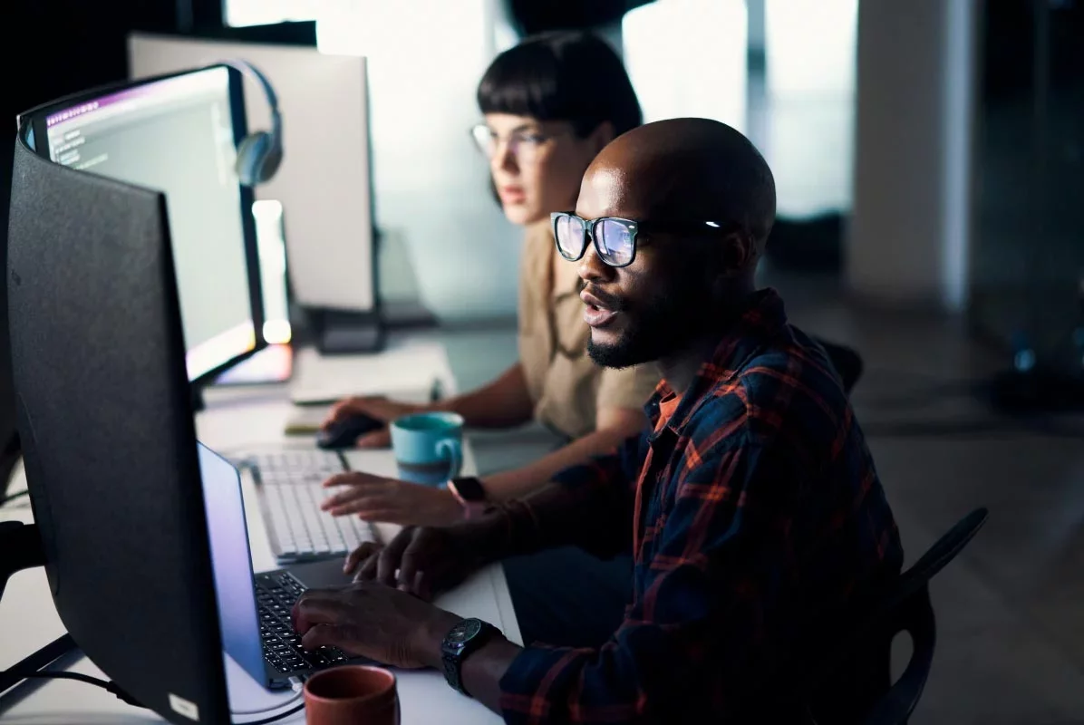 Two cybersecurity engineers working side by side at a workstation, reviewing code and system activity on large monitors—representing Proofpoint’s secure connectivity layer for enterprise AI.
